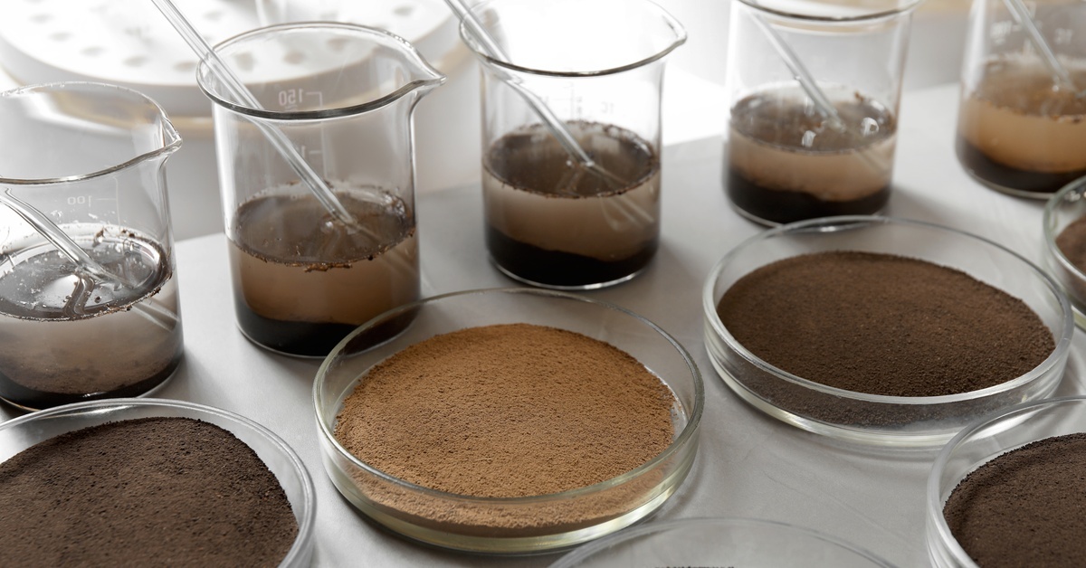 Several petri dishes and beakers filled with various colors and textures of soil in a laboratory environment.