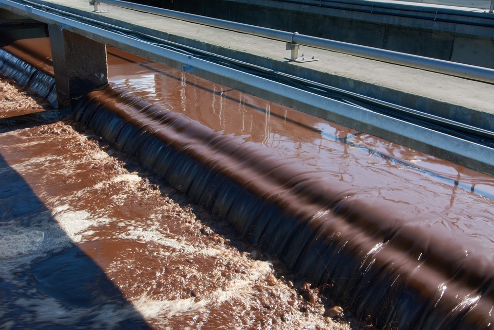 Activated sludge overflows a concrete weir as the brownish water flows steadily through the treatment structure.