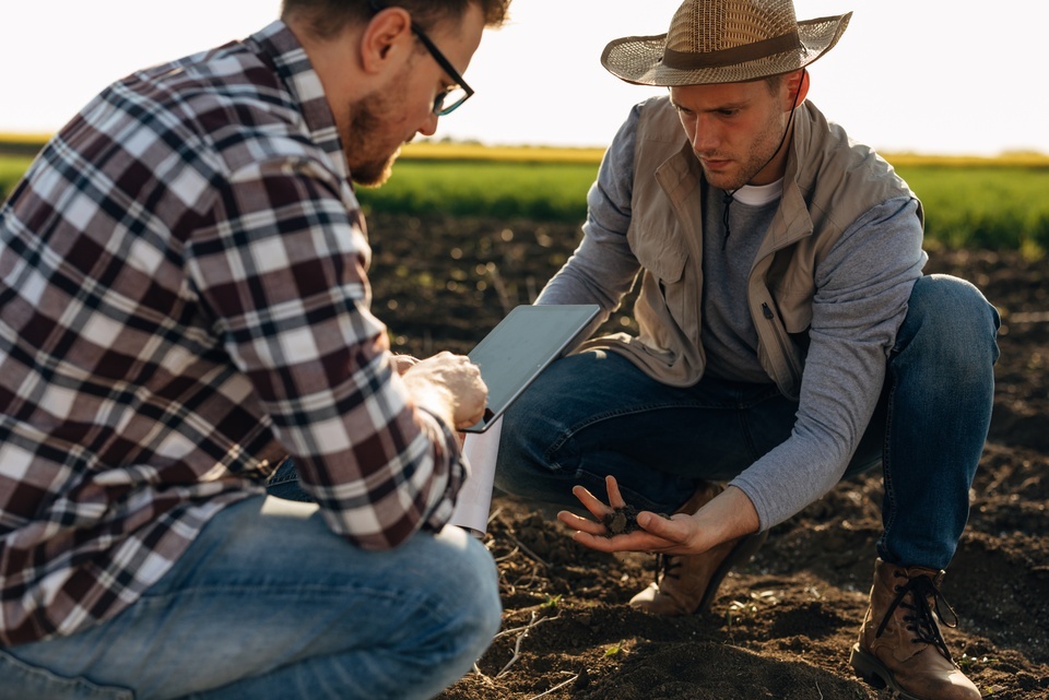 Two men crouch on a stretch of dirt. One holds a scoop of soil while the other looks down at a tablet.