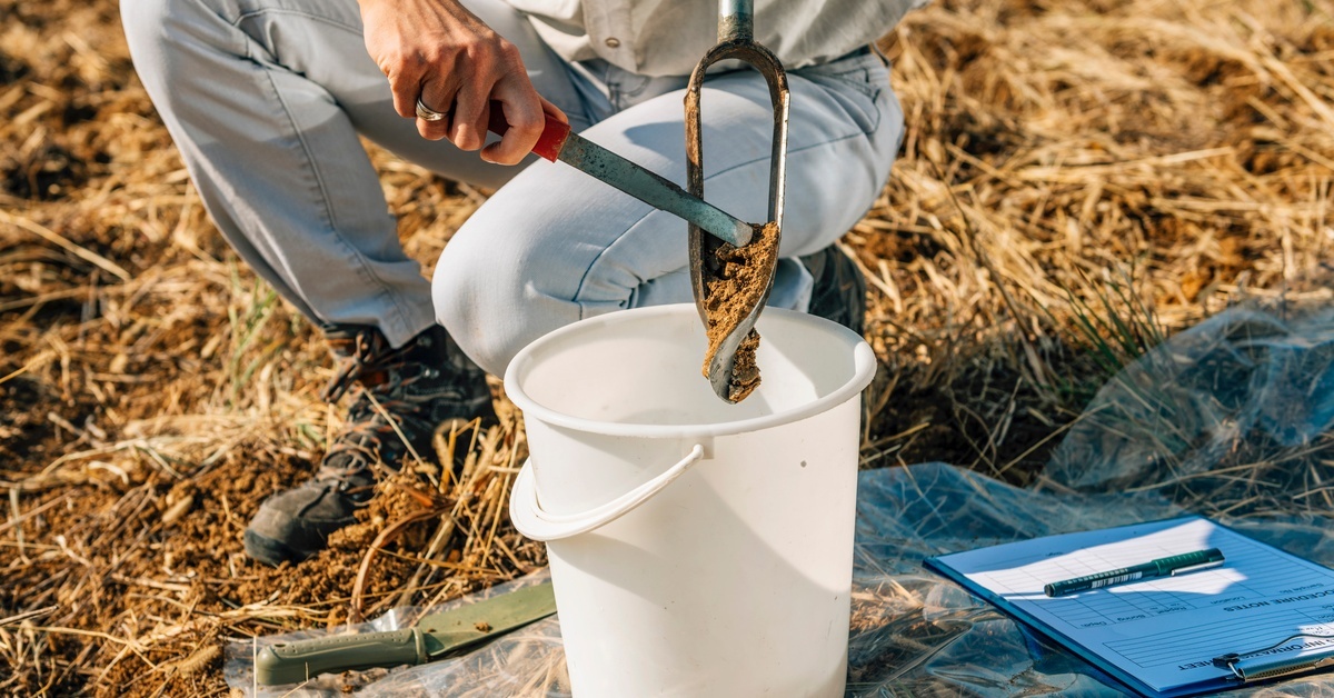 A person scrapes a soil sample with a tool into a bucket with a shovel, clipboard, and plastic sheet in view.