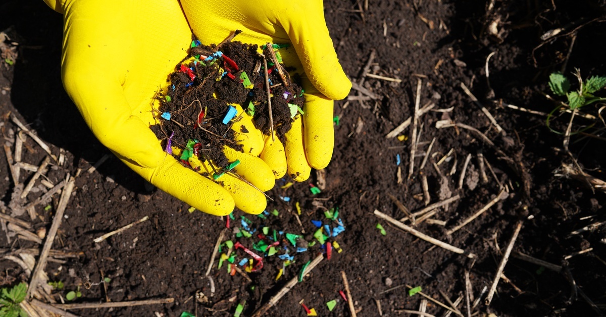 Gloved hands hold soil mixed with small pieces of colorful plastic. The soil below also contains colorful bits of plastic.