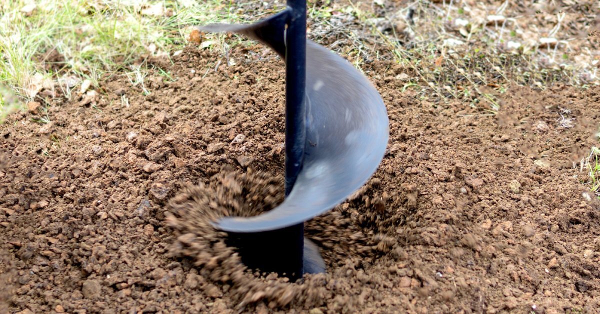 A drill with a long, black auger bit rotates in soil, sending loose dirt into the air during operation.
