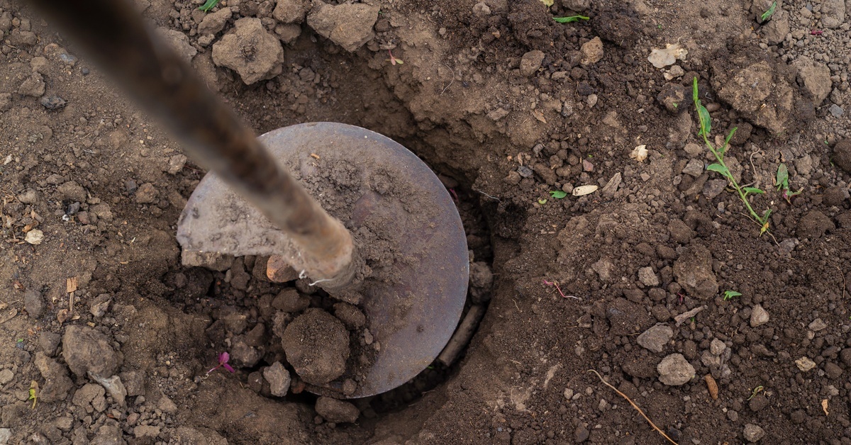 A spiral auger tool is partially embedded in dark soil, showing loose dirt around the drilling area.