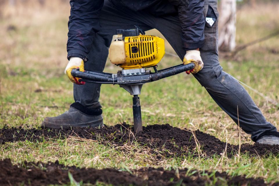A person wearing gloves operates a yellow gas-powered drill with a black auger, displacing soil in a grassy yard.