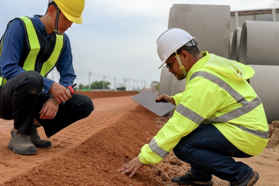 Two men wearing hard hats crouch on laterite soil. One man holds a notebook while touching the soil with his other hand.