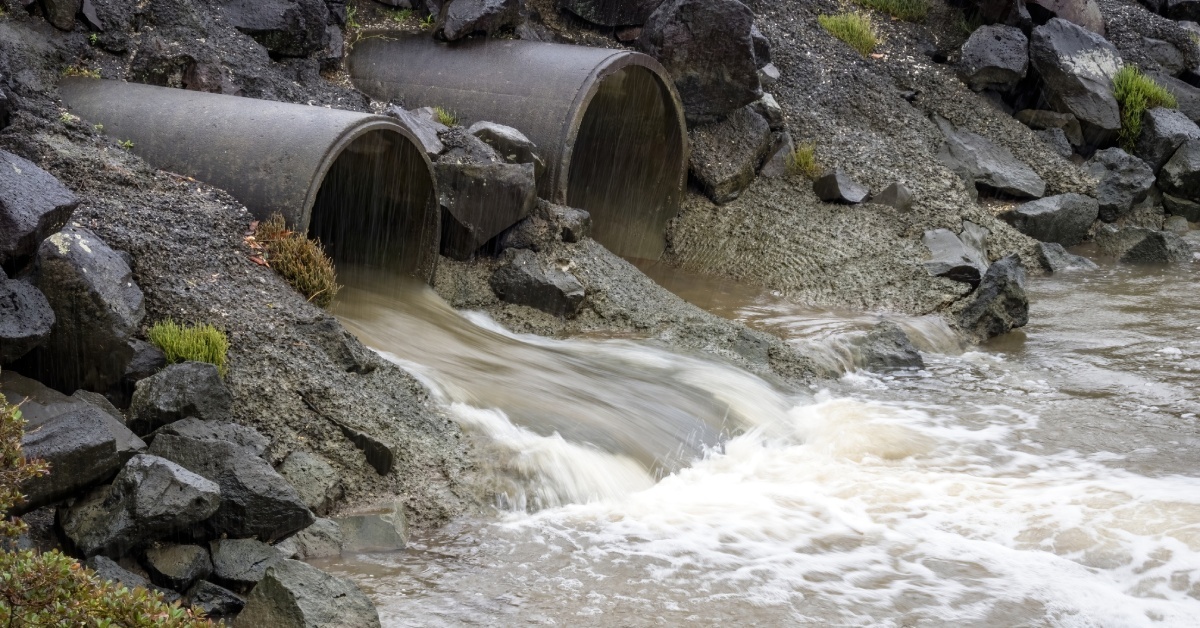 Brownish water flowing from two concrete pipes. There are multiple large rocks surrounding the pipes.