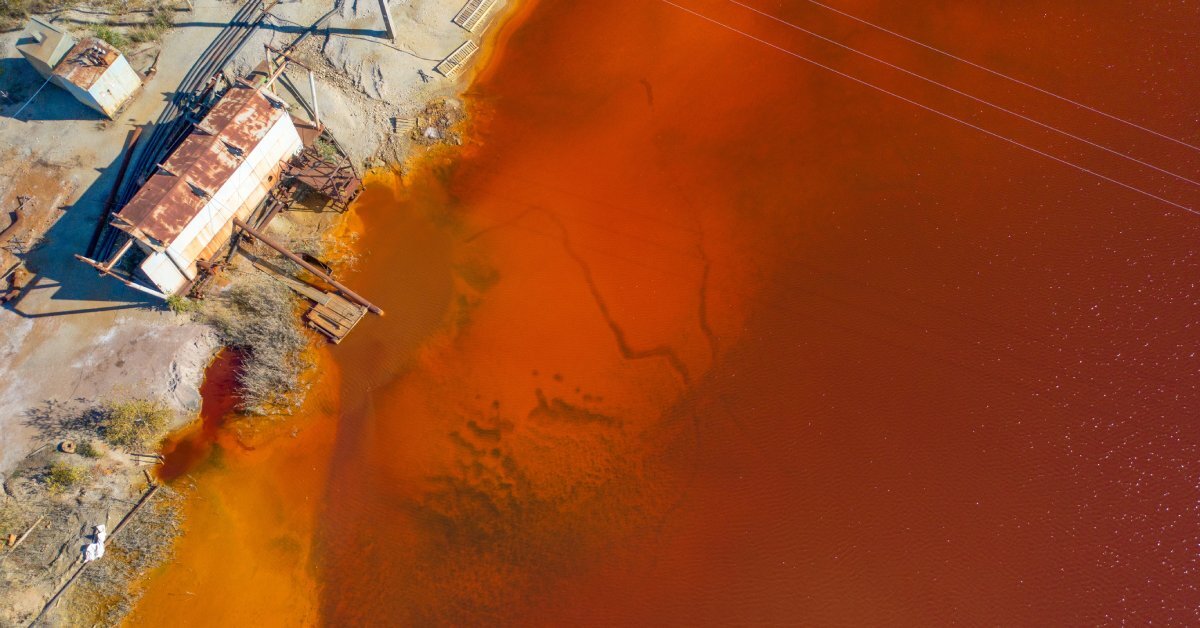 Rust-colored water surrounds a small shore building with a rusted roof and a dock extending outward.