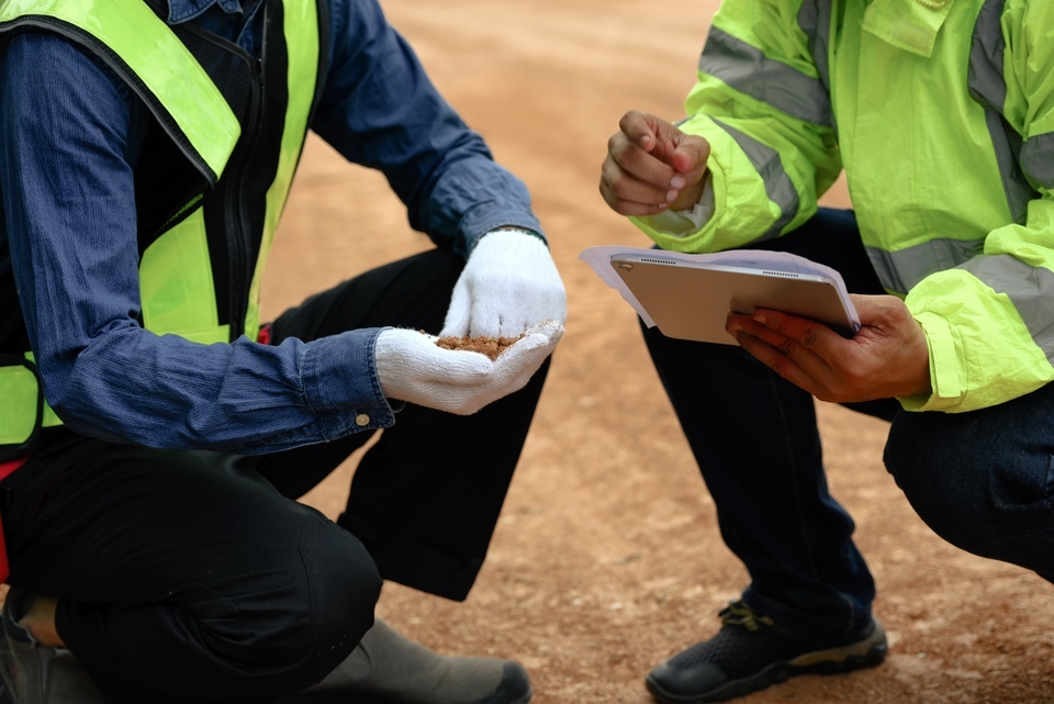 A man in gloves swirls dirt in his hand, while another holds a tablet, partly covered by a piece of paper.