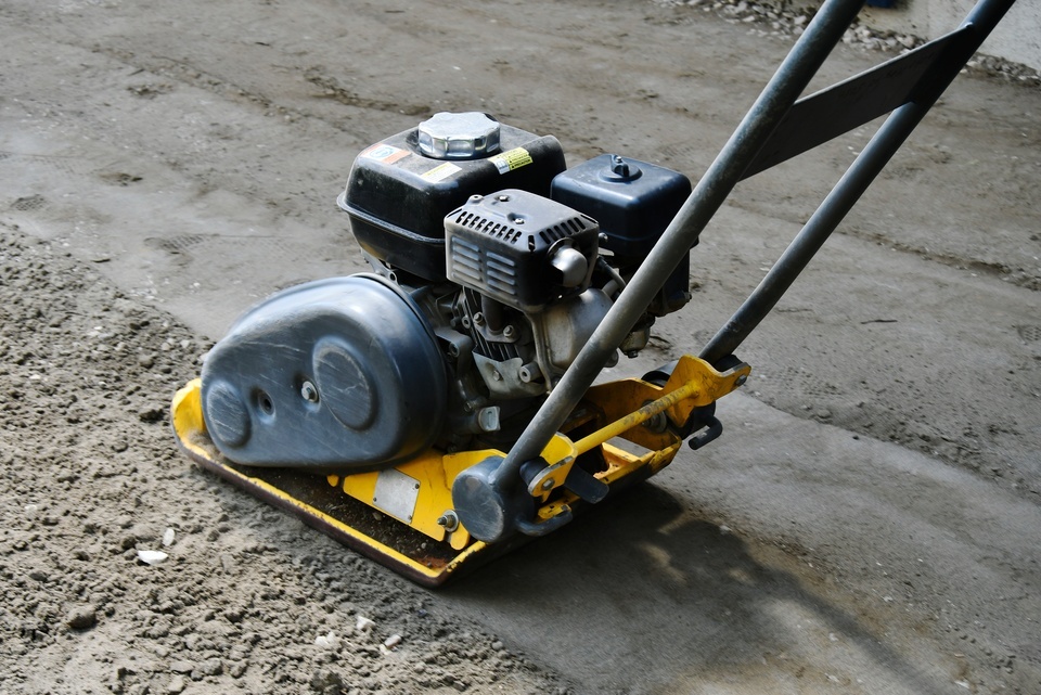 A yellow soil compactor rolls over greige sandy soil mixed with gravel, flattening the ground's surface.