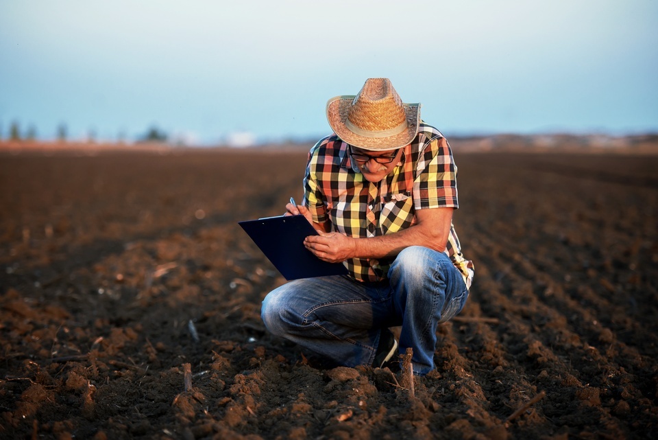 A man in glasses and a plaid shirt squats over a plowed field while writing on a clipboard under a pale blue sky.