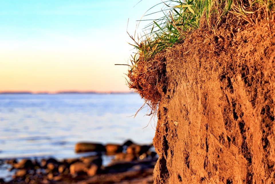 A close-up of a reddish-brown cliff shows green grass on top, with calm blue water under a golden sky in the background.