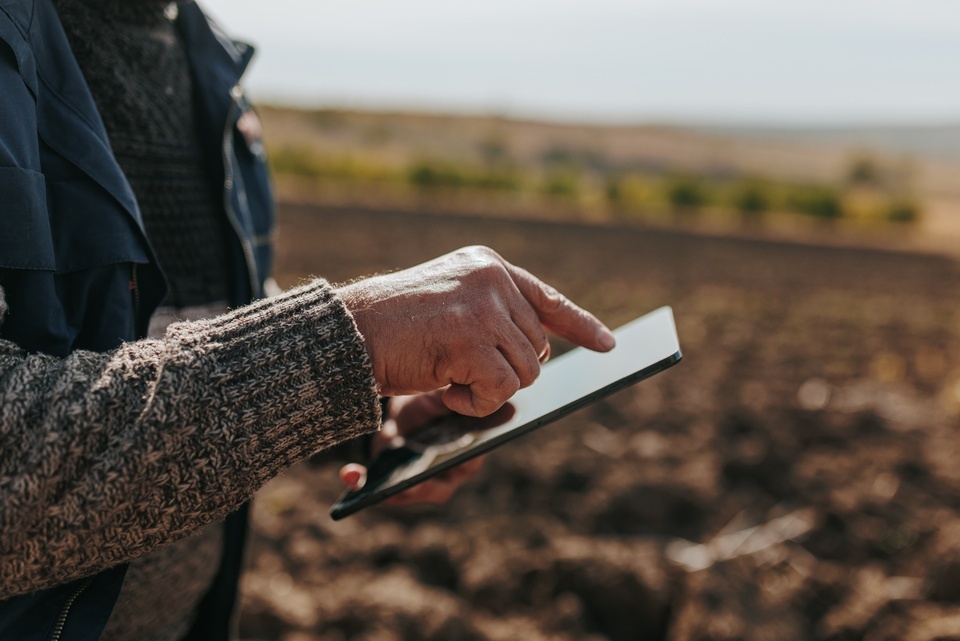A person in a navy jacket uses a tablet in a tilled field, with blurred green bushes and an overcast sky in the background.