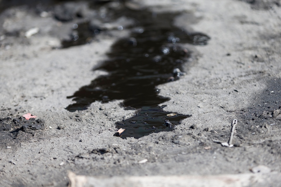 A dark oil puddle snakes across dusty concrete, reflecting light near a cigarette butt and a small metal nail.