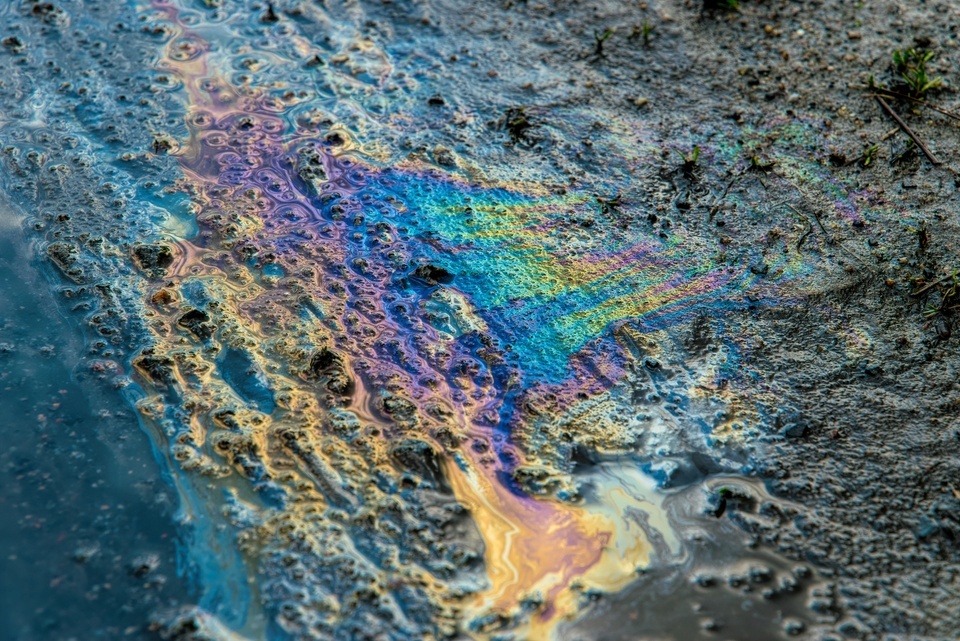 A rainbow oil sheen spreads across muddy ground, shimmering over pebbles, grit, and sparse grass after a spill.