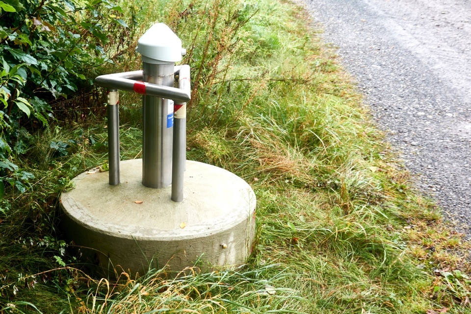 A metal groundwater well rises from a concrete base as tall grass and a gravel road surround the site.