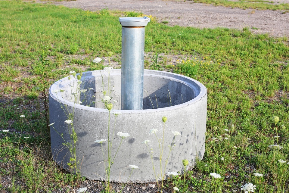 A steel monitoring well stands inside a concrete ring while wildflowers and grass cover the open field.
